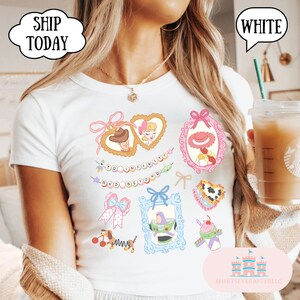 Op de afbeelding: Wit T-shirt met kleurrijke cartoonafbeeldingen van personages en elementen uit een populaire animatiefilm. Het shirt heeft ingelijste afbeeldingen, strikken en de tekst "TO INFINITY AND BEYOND".