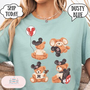 May include: A dusty blue t-shirt featuring a graphic print of four brown teddy bears wearing black Mickey Mouse ear hats. One bear holds a red Mickey-shaped balloon, another wears a white shirt with an M, and one carries a red and black backpack.