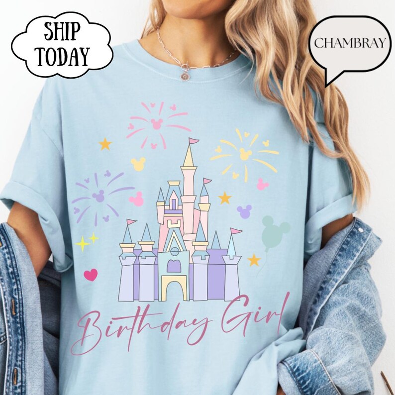 May include: Pale blue t-shirt with a castle graphic, fireworks, stars, and Mickey Mouse silhouettes. The text "Birthday Girl" is in a script font. The shirt has a relaxed fit.