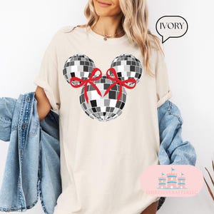 May include: A white t-shirt featuring a black and white disco ball Mickey Mouse design. The disco balls have red bows. The shirt is labelled "IVORY".