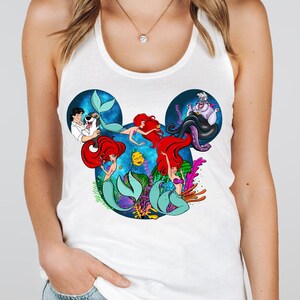 Puede incluir: Camiseta sin mangas blanca con un gr&aacute;fico colorido de Ariel y otros personajes de La Sirenita. El dise&ntilde;o tiene forma de orejas de Mickey Mouse, con Ariel y otros personajes en una escena marina.