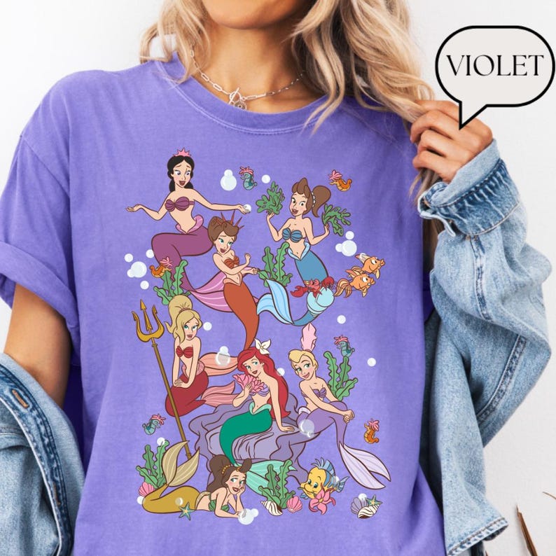 Op de afbeelding: Een violetkleurig T-shirt met een kleurrijke illustratie van verschillende zeemeerminnen. De zeemeerminnen worden afgebeeld in verschillende poses met verschillende haarkleuren en zeemeerminstaarten. Het woord "VIOLET" staat in een tekstballon rechtsboven.