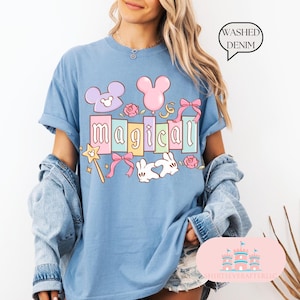 Op de afbeelding: Lichtblauw T-shirt met het woord "magical" in pastelkleuren, samen met cartoonafbeeldingen van een muis, een ballon, strikken, rozen, een toverstaf en twee witte konijnen. De tekst "Washed Denim" is ook aanwezig.