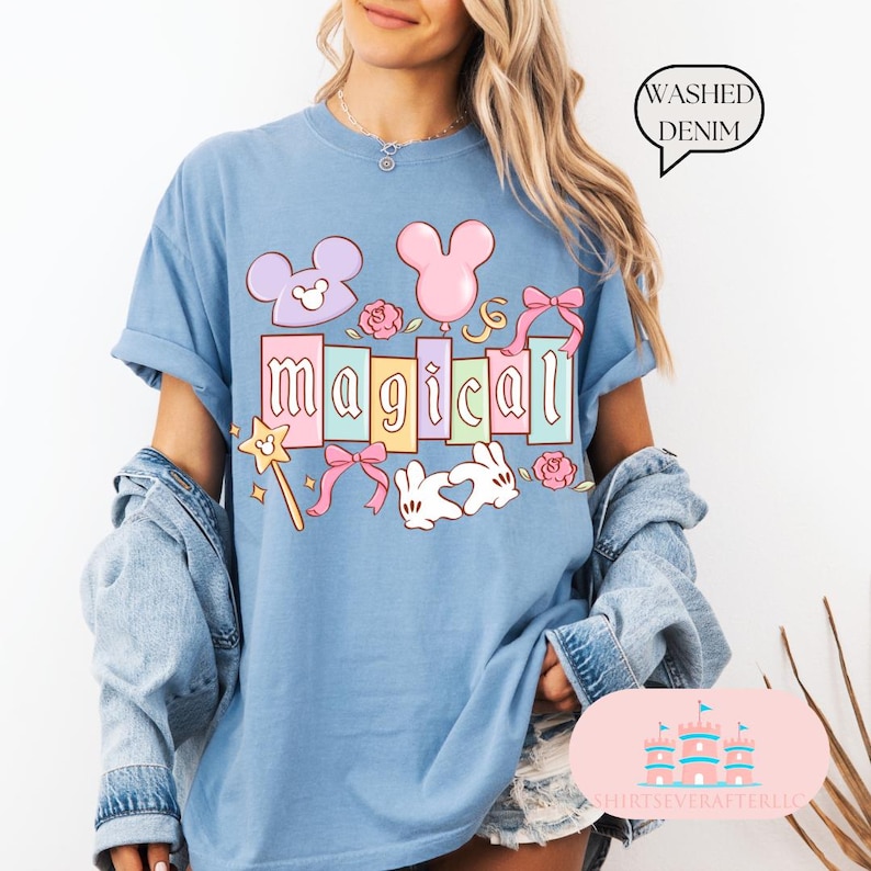 Op de afbeelding: Lichtblauw T-shirt met het woord "magical" in pastelkleuren, samen met cartoonafbeeldingen van een muis, een ballon, strikken, rozen, een toverstaf en twee witte konijnen. De tekst "Washed Denim" is ook aanwezig.