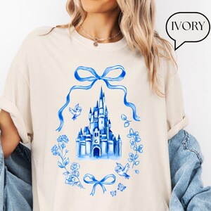 以下が含まれることがあります： アイボリー色のTシャツ。青い水彩画の城のデザイン、リボン、鳥、花の要素が特徴です。「IVORY」の文字が吹き出しに入っています。デザインはシャツの中央に配置されています。
