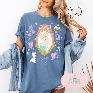 K&ouml;nnte beinhalten: Ein blaues Jeans-T-Shirt mit einem Blumendesign, das ein gerahmtes Bild von Elsa aus Frozen zeigt. Der Rahmen ist golden mit einem rosa Band und Schleife. Der Text "BLUE JEAN" befindet sich in einem wei&szlig;en Kasten in der oberen rechten Ecke des Bildes.