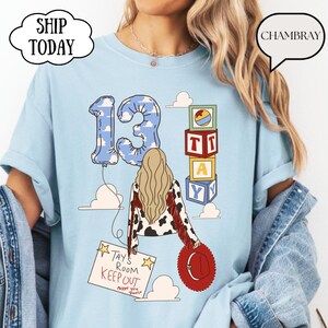 May include: A light blue t-shirt featuring a graphic of a person with long blonde hair, a cow-print jacket with red fringe, and a red hat. The design includes a blue cloud-patterned number 13 balloon, alphabet blocks, and a sign reading TAY'S ROOM KEEP OUT.