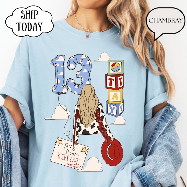 May include: A light blue t-shirt featuring a graphic of a person with long blonde hair, a cow-print jacket with red fringe, and a red hat. The design includes a blue cloud-patterned number 13 balloon, alphabet blocks, and a sign reading TAY'S ROOM KEEP OUT.