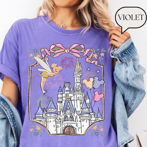 Puede incluir: Camiseta violeta con una ilustraci&oacute;n de cuento de hadas. El dise&ntilde;o incluye un hada, fuegos artificiales y un castillo, con un lazo rosa y globos de Mickey Mouse. La palabra "Violet" est&aacute; en una burbuja. El tema general es un cuento de hadas.