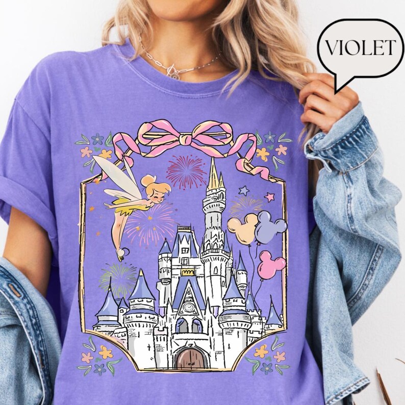 Puede incluir: Camiseta violeta con una ilustraci&oacute;n de cuento de hadas. El dise&ntilde;o incluye un hada, fuegos artificiales y un castillo, con un lazo rosa y globos de Mickey Mouse. La palabra "Violet" est&aacute; en una burbuja. El tema general es un cuento de hadas.