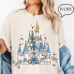 May include: Ivory-colored t-shirt featuring a detailed illustration of a fairytale castle with blue turrets and gold accents. The words "DISNEY WORLD" are written in a whimsical font. Black stars are scattered around the castle design.