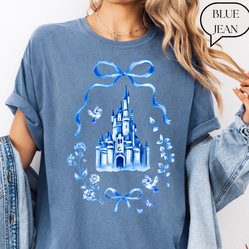 以下が含まれることがあります： 青いジーンズ色のTシャツ。青いお城のイラストが、花のリース、リボン、鳥に囲まれています。「BLUE JEAN」の文字が吹き出しに入っています。シャツの袖はロールアップされています。