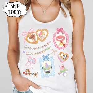 Op de afbeelding: Witte tanktop met kleurrijke cartoon illustraties van personages en elementen uit een populaire animatiefilm. Het ontwerp bevat ingelijste portretten, strikken en de zin "TO INFINITY AND BEYOND".