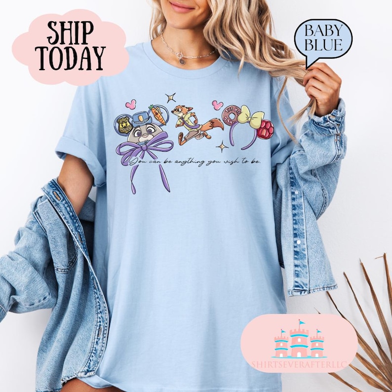 May include: Light blue t-shirt featuring cartoon character designs and the text "You can be anything you wish to be." The shirt has a crew neck and short sleeves. The design includes a bunny, fox, and donut-shaped headbands.