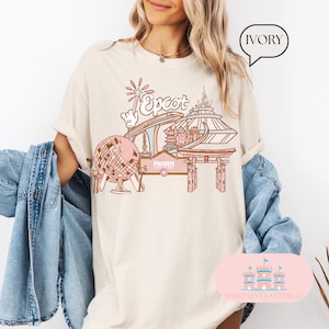 May include: A white t-shirt featuring a pink and blue illustration of Epcot, a theme park at Walt Disney World. The illustration includes the Spaceship Earth ride, the World Showcase, and the Frozen Ever After ride. The text "Epcot" is written in pink letters on the shirt.
