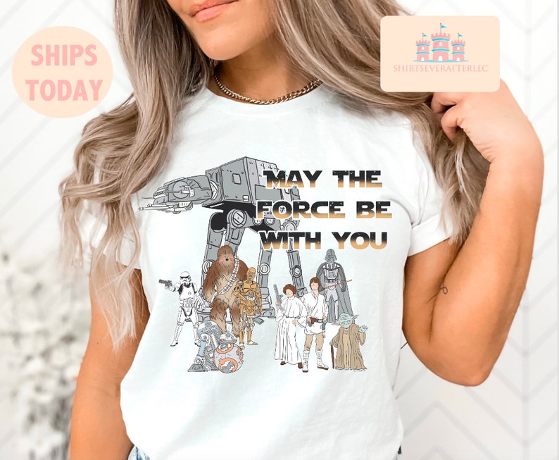 Puede incluir: Camiseta blanca con un gr&aacute;fico de personajes de Star Wars, incluyendo Darth Vader, Chewbacca, R2-D2 y un caminante AT-AT. El texto "Que la Fuerza te acompa&ntilde;e" est&aacute; impreso en dorado en la camiseta.