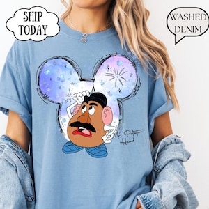 Può includere: Una t-shirt blu denim slavato con una grafica di Mr. Potato Head centrata all'interno di una silhouette delle orecchie di Topolino in stile acquerello. Il design include stelle, fuochi d'artificio e il testo "Mr. Potato Head" in corsivo nero.