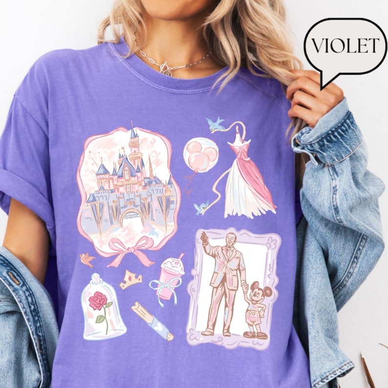 Op de afbeelding: Een violetkleurig T-shirt met diverse illustraties, waaronder een kasteel, een jurk en een ingelijste afbeelding van een man en een muis. Het woord "VIOLET" staat in een tekstballon. De illustraties zijn in tinten roze, blauw en wit.