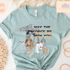 Puede incluir: Una camiseta azul claro con un estampado gr&aacute;fico de personajes de Star Wars, incluyendo Darth Vader, Chewbacca, R2-D2 y un caminante AT-AT. El texto "Que la Fuerza te acompa&ntilde;e" est&aacute; impreso en la camiseta.
