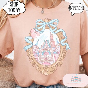 Puede incluir: Camiseta color melocot&oacute;n con un dise&ntilde;o de espejo con marco dorado que muestra un castillo, un lazo azul claro y detalles en rosa. La camiseta tiene el texto "(H/PEACH)" y "SHIP TODAY".