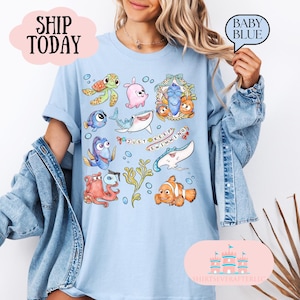 May include: Light blue t-shirt featuring cartoon sea creatures and the phrase "Just Keep Swimming." The design includes a turtle, shark, clownfish, and other characters. The shirt is a baby blue color.