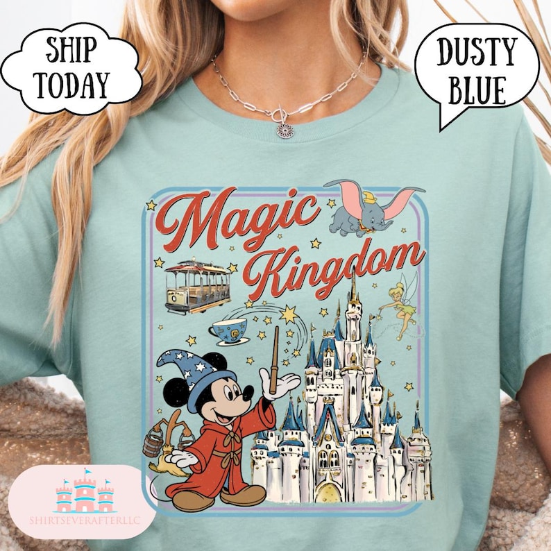 May include: A light blue t-shirt with a graphic featuring Mickey Mouse as a wizard, the words "Magic Kingdom", and images of Dumbo, a castle, and other Disney-themed elements. The shirt has a crew neck and short sleeves.
