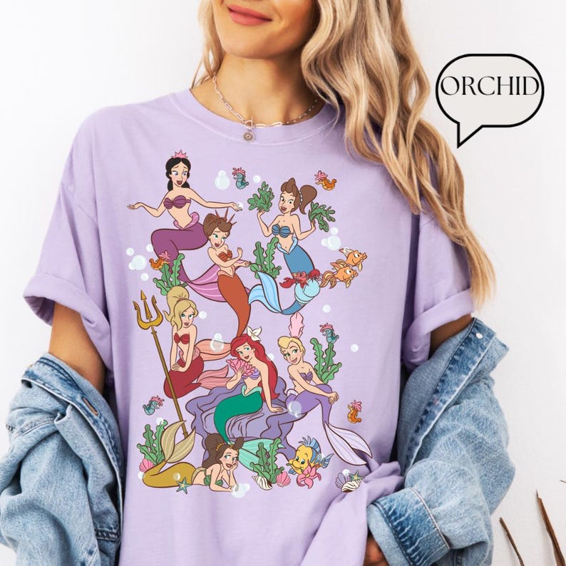Op de afbeelding: Een lichtpaars T-shirt met een kleurrijke illustratie van zeemeerminnen en zeedieren. Het ontwerp toont diverse zeemeerminnen met verschillende haarkleuren en outfits, omringd door zeewier, vissen en bellen. Het woord "ORCHID" staat in een tekstballon.