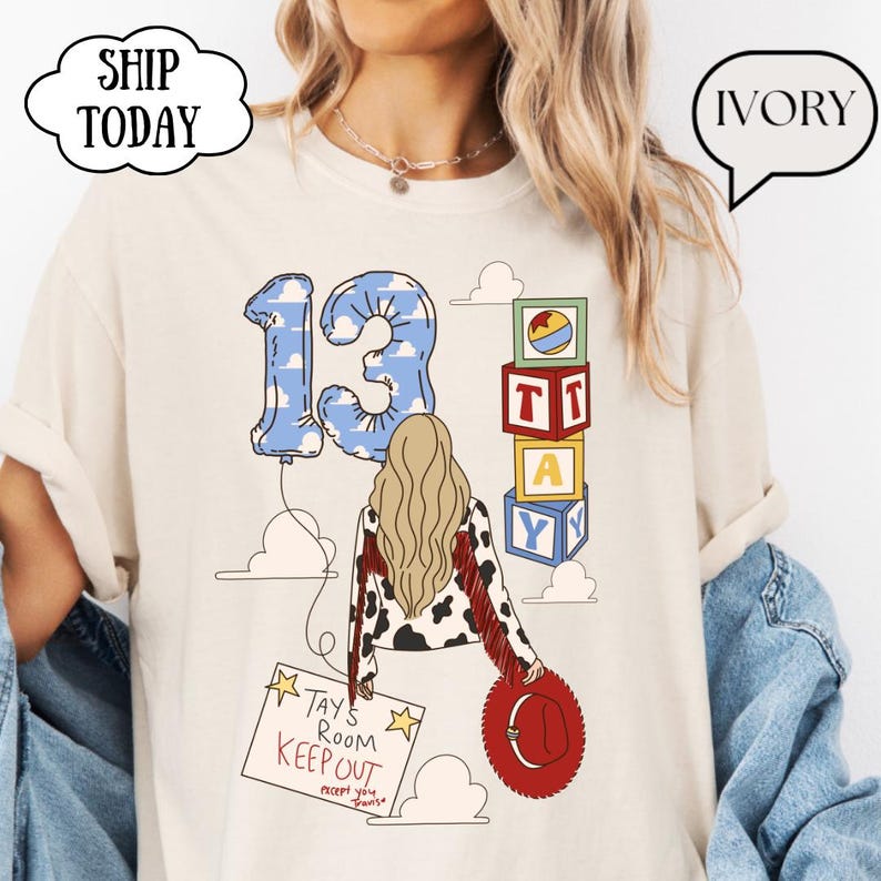 May include: An ivory t-shirt featuring a graphic of a person with long blonde hair, a cow-print jacket with red fringe, and a red hat. The design includes a blue cloud-patterned number 13 balloon, alphabet blocks, and a sign reading TAY'S ROOM KEEPOUT.