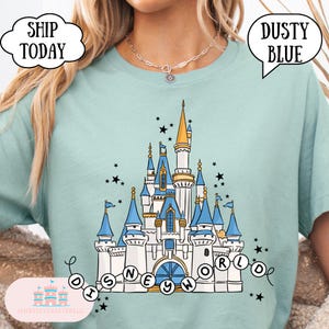 May include: Dusty blue t-shirt featuring a detailed illustration of a castle with blue and gold accents, surrounded by stars. The text "DISNEYWORLD" is displayed below the castle. The shirt also has the text "SHIP TODAY" and "DUSTY BLUE".