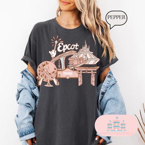May include: A dark grey t-shirt featuring a graphic of Epcot, a popular theme park in Orlando, Florida. The graphic depicts a stylised illustration of the park's iconic Spaceship Earth, the World Showcase Lagoon, and the Frozen Ever After ride. The text "Epcot" is printed in a pink font.