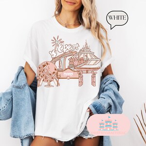 May include: White t-shirt featuring a pink and brown illustration of Epcot, including the Spaceship Earth ride, the Japan pavilion, and the Frozen Ever After ride.