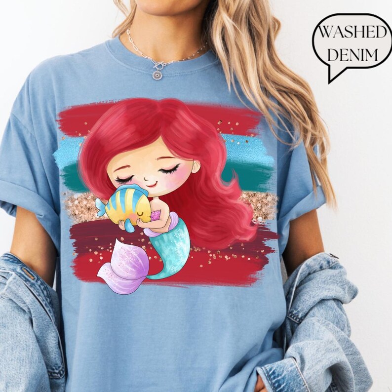 Puede incluir: Camiseta azul claro con un dise&ntilde;o de sirena de dibujos animados. La sirena tiene el pelo rojo y una cola turquesa, sosteniendo un pez amarillo y azul. El gr&aacute;fico est&aacute; sobre pinceladas rojas, azules y doradas. El texto dice "WASHED DENIM".