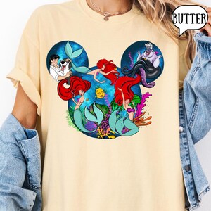 Puede incluir: Camiseta amarillo claro con un dise&ntilde;o de los personajes de La Sirenita de Disney dispuestos en forma de cabeza de Mickey Mouse. El dise&ntilde;o incluye a Ariel, el Pr&iacute;ncipe Eric, &Uacute;rsula y Flounder.