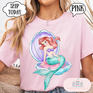 May include: Pale pink t-shirt with a mermaid illustration in a seashell. The mermaid has red hair and a teal tail. The shirt has the text "PINK" and "SHIP TODAY".