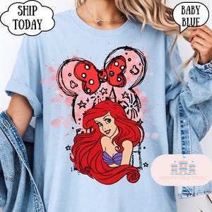 May include: Baby blue t-shirt featuring a graphic of Ariel from The Little Mermaid, with a red bow and Mickey Mouse ears design. The shirt has the text "Ship Today" and "Baby Blue". The shirt is from ShirtsEverAfterLLC.