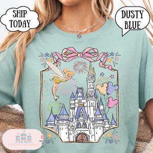 Puede incluir: Camiseta azul polvoriento con una ilustraci&oacute;n caprichosa de un hada, fuegos artificiales y un castillo. El dise&ntilde;o incluye un lazo rosa, globos y el texto "Dusty Blue" en una burbuja. La camiseta tambi&eacute;n tiene el texto "Ship Today" en una nube.