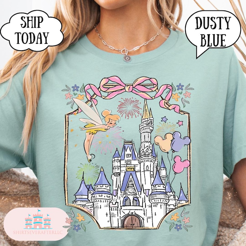 Puede incluir: Camiseta azul polvoriento con una ilustraci&oacute;n caprichosa de un hada, fuegos artificiales y un castillo. El dise&ntilde;o incluye un lazo rosa, globos y el texto "Dusty Blue" en una burbuja. La camiseta tambi&eacute;n tiene el texto "Ship Today" en una nube.
