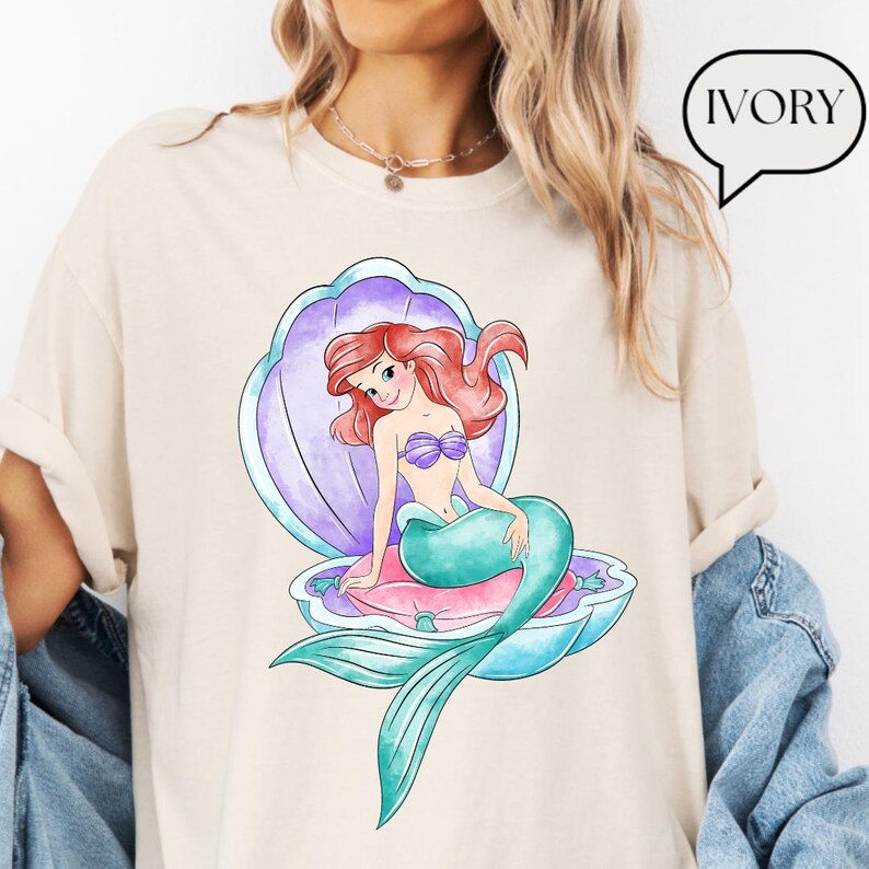 May include: Ivory-coloured t-shirt featuring a cartoon mermaid with red hair, a purple seashell, and a teal tail. The mermaid is seated on a pink pillow inside the shell. The word "IVORY" is in a speech bubble.