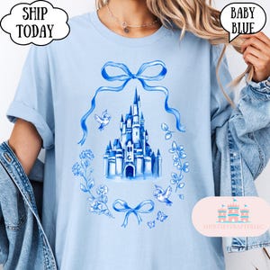 以下が含まれることがあります： 水彩画の城のイラストが特徴のベビーブルーのTシャツ。青色の花輪とリボンで囲まれています。「SHIP TODAY」と「BABY BLUE」の文字も表示されています。袖は折り返されています。