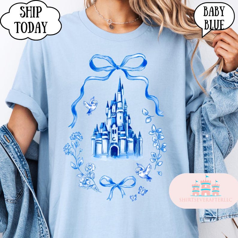 以下が含まれることがあります： 水彩画の城のイラストが特徴のベビーブルーのTシャツ。青色の花輪とリボンで囲まれています。「SHIP TODAY」と「BABY BLUE」の文字も表示されています。袖は折り返されています。