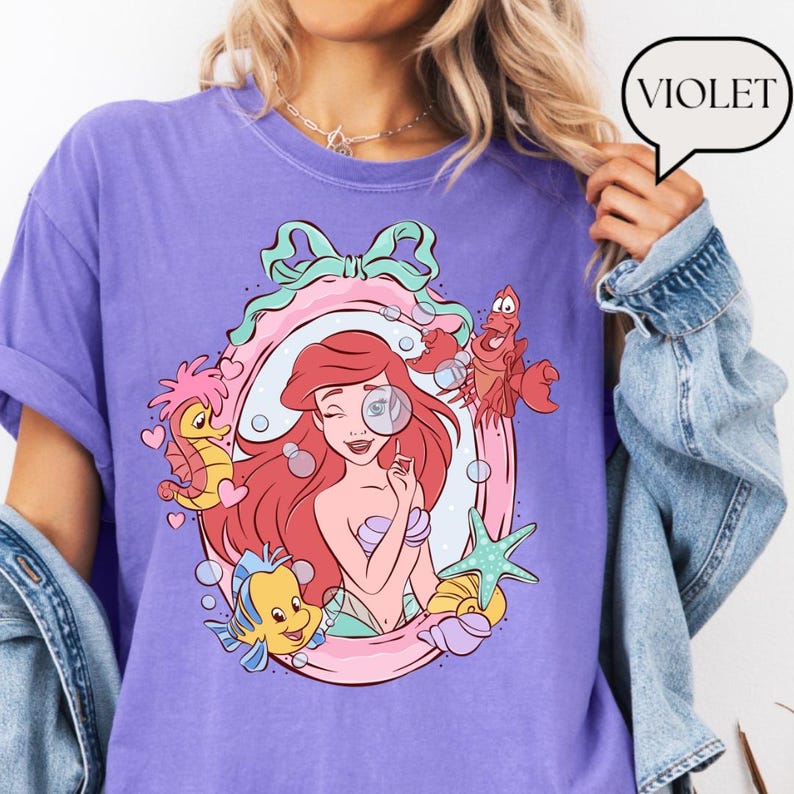 May include: Violet-colored t-shirt featuring a cartoon illustration of a mermaid with red hair, surrounded by sea creatures. The design includes a pink and green frame, bubbles, and the word "VIOLET" in a speech bubble.