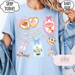 Op de afbeelding: Babyblauwe t-shirt met diverse cartoonkarakters. Het shirt heeft ingelijste afbeeldingen van personages, strikken en de tekst "TO INFINITY AND BEYOND". Het shirt heeft ook de tekst "SHIP TODAY" en "BABY BLUE".
