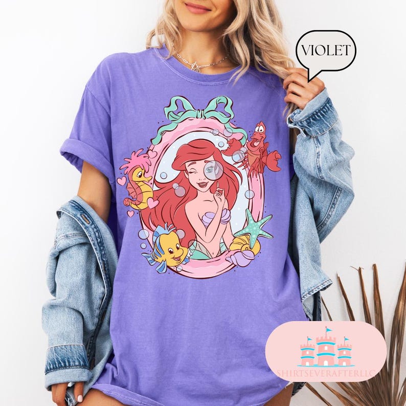 K&ouml;nnte beinhalten: Violettes T-Shirt mit einer Cartoon-Illustration von Arielle, der Meerjungfrau. Das Design umfasst Arielle, Fabius, Sebastian und andere Meereskreaturen in einem dekorativen Rahmen. Das Shirt hat eine lockere Passform und kurze &Auml;rmel.