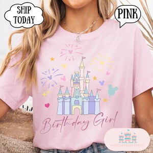 May include: Pale pink t-shirt with a castle design, fireworks, stars, and Mickey Mouse shapes. The words "Birthday Girl" are written in cursive. The shirt also has the text "Pink" in a speech bubble.