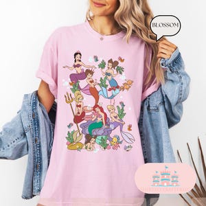 May include: Pink t-shirt featuring a colorful illustration of mermaids in a sea scene. The graphic includes various mermaids with different hair colors and outfits, surrounded by seaweed, bubbles, and fish. The shirt has the word "BLOSSOM" in a speech bubble.