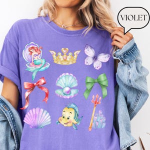Może przedstawiać: Fioletowa koszulka z akwarelowymi ilustracjami syreny, korony, motyla, kokardy, muszli, perły, ryby, tr&oacute;jzębu i koralowc&oacute;w. Słowo "VIOLET" znajduje się w dymku. Styl casual.