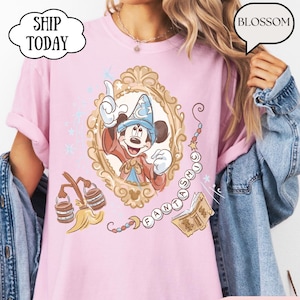K&ouml;nnte beinhalten: Hellrosa T-Shirt mit Mickey Mouse als Zauberer in einem goldenen Rahmen. Das Design enth&auml;lt das Wort "FANTASMIC" und andere magische Elemente. Das Shirt hat den Text "BLOSSOM" in einer Sprechblase.