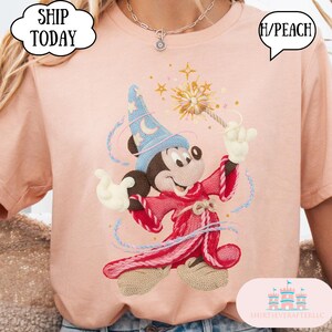 May include: Peach-coloured t-shirt with an embroidered design of Mickey Mouse as a wizard, holding a magic wand. Mickey wears a blue wizard hat with a crescent moon and stars. The shirt has the text "SHIP TODAY" in a white cloud-shaped bubble.