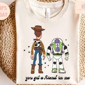 May include: A white t-shirt featuring a cartoon image of Woody and Buzz Lightyear from the Toy Story film. The characters are facing away from the viewer. The text "you got a friend in me" is printed below the characters.