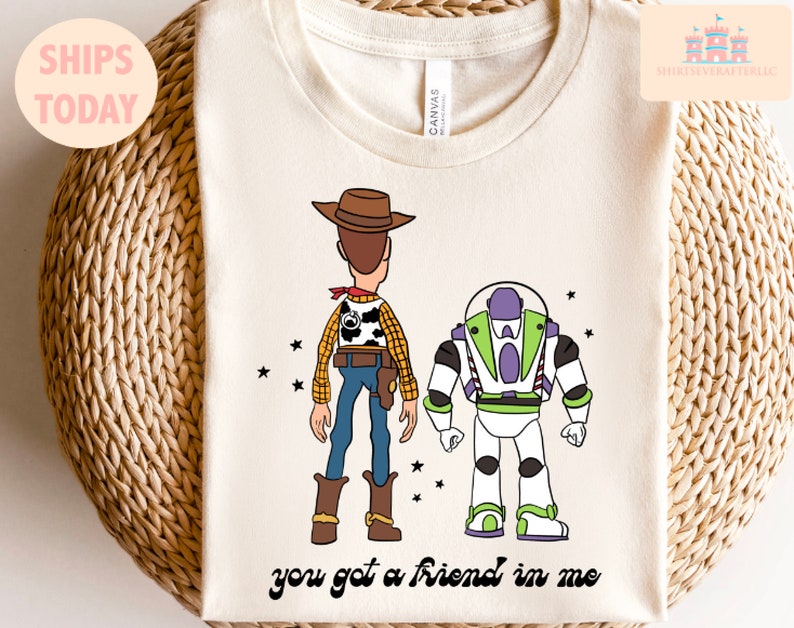 May include: A white t-shirt featuring a cartoon image of Woody and Buzz Lightyear from the Toy Story film. The characters are facing away from the viewer. The text "you got a friend in me" is printed below the characters.